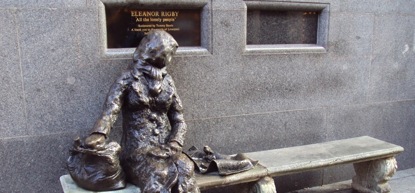 a metal statue of a faceless woman in a coat and headscarf sat on a bench outside with a plaque that reads "eleanor rigby 'all the lonely people'"